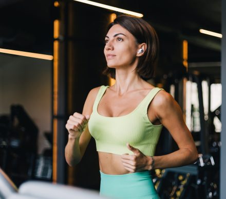 Fit caucasian beautiful woman in fitting sport wear at gym running jogging at treadmill with wireless headphones in ears